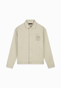 Beige katoenen jack met een kraag, drukknoopsluiting, zijzakken en een geborduurd logo op de borstzak. Een gestructureerd weefpatroon is zichtbaar.