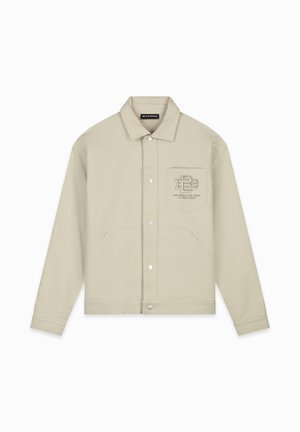 Beige katoenen jack met een kraag, drukknoopsluiting, zijzakken en een geborduurd logo op de borstzak. Een gestructureerd weefpatroon is zichtbaar.