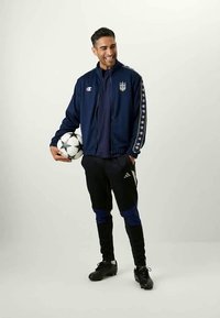 Un homme portant une veste de sport bleu marine et un pantalon noir Adidas se tient en souriant, tenant un ballon de football blanc et noir à motifs.