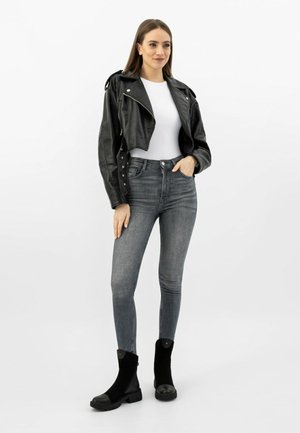Young woman standing in black cropped leather jacket, white top, grey skinny jeans, and black ankle boots against plain white background.