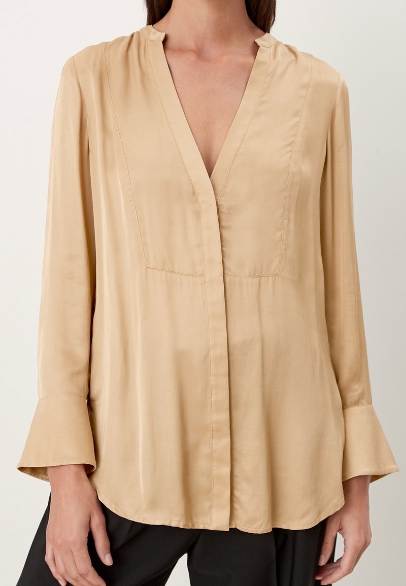 Blouse en soie beige avec un décolleté en V, des manches longues évasées et des détails de couture subtils. Texture lisse avec une coupe décontractée.