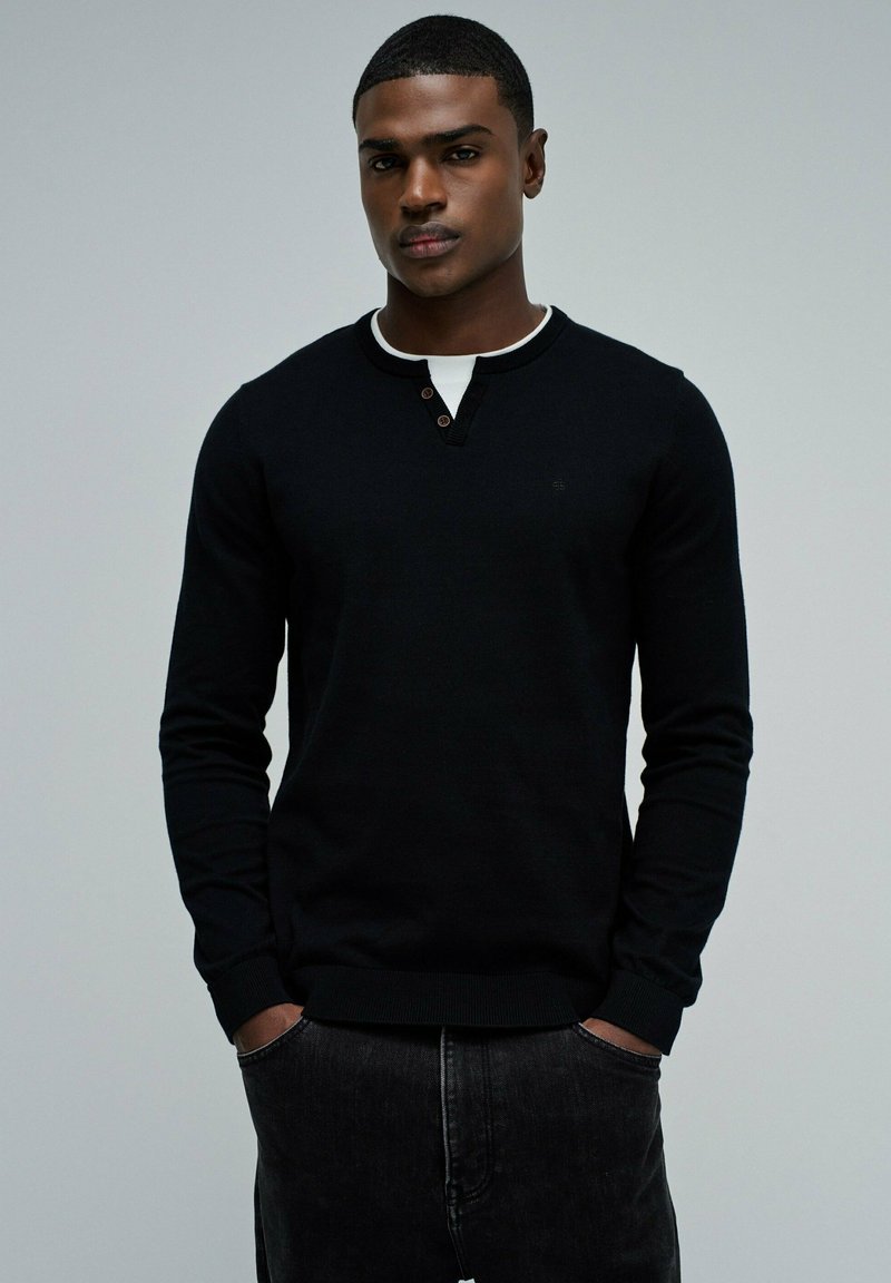 Suéter negro de manga larga con cuello redondo, que presenta un interior blanco en el cuello y detalles de botones. Combinado con jeans de mezclilla oscuros.