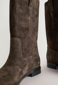 Bottes hautes en suède marron, présentant une texture douce, une silhouette élancée, et des talons en bloc noirs bas avec des détails de couture subtils.