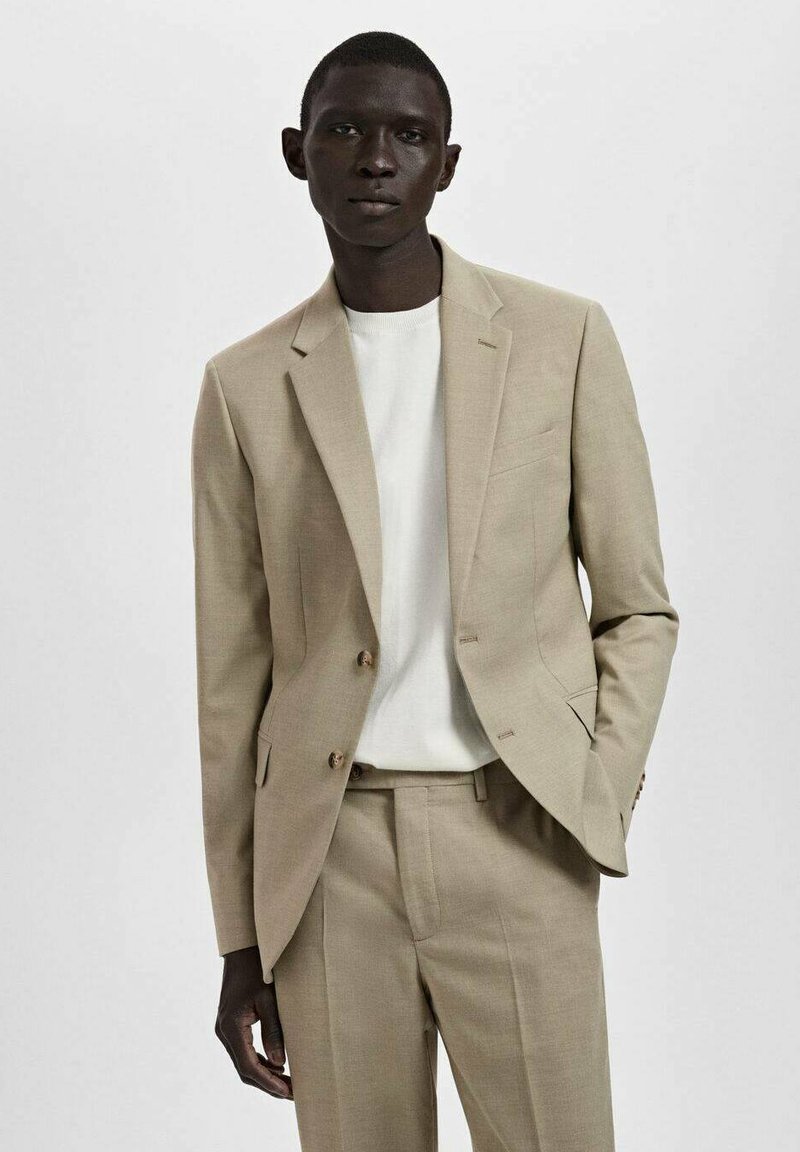 Completo beige con giacca su misura e pantaloni con tasche piatte, abbinato a una camicia bianca a girocollo. Tessuto liscio, due bottoni, cuciture minime.