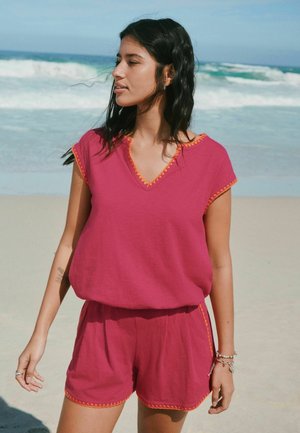 Woman with black hair wearing a sleeveless magenta top and matching shorts with orange trim standing on a sandy beach by the ocean.