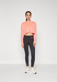 Langermet crop-top i lys korall med lagdelt design og logo. Tette svarte leggings med ankelglidelåser og en glatt tekstur. Hvite joggesko.