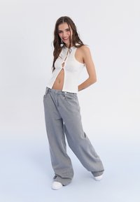 Top corto blanco de canalé con un diseño de ojal, combinado con pantalones de mezclilla gris holgados y zapatillas blancas sobre un fondo claro.