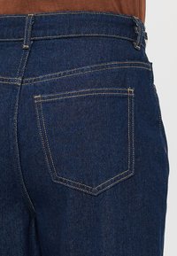 Jean en denim bleu foncé avec une texture lisse, doté d'une seule poche arrière et de coutures contrastées, conçu avec une taille mi-haute.