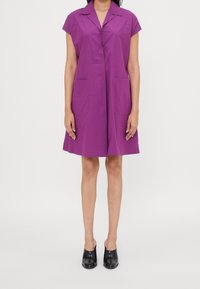 Vestito corto viola a maniche corte con colletto, due tasche frontali e una vestibilità morbida, abbinato a scarpe aperte nere.