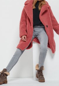 Różowy płaszcz ze sztucznego kożucha z głębokim kołnierzem, o luźnym kroju, zapinany na guziki, zestawiony z szarymi obcisłymi dżinsami i brązowymi butami wyłożonymi futerkiem.