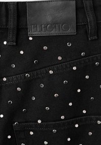 Sección de tela de mezclilla negra con un bolsillo trasero, adornada con pequeños tachuelas plateadas dispersas y una etiqueta de cuero negro en la cintura.