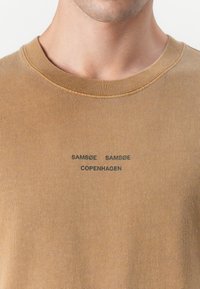 Sudadera de algodón marrón con cuello redondo y ribete en el cuello. Presenta el texto impreso "SAMSØE SAMSØE COPENHAGUE" en negro. Diseño simple y limpio.