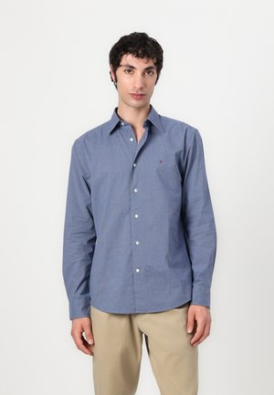 Blue patterned button-up shirt with long sleeves, featuring white buttons and a small logo on the chest pocket, paired with beige pants.