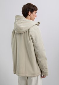 Young person standing sideways wearing a beige hooded jacket and off-white pants against a plain light background.