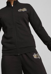 Chaqueta de chándal negra con cremallera y puños y dobladillo acanalados, que presenta bordados florales en el pecho en acentos dorados y blancos.