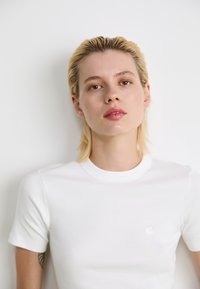 Ung person med kort blondt hår iført en hvid ribbet t-shirt mod en ensfarvet hvid baggrund, som ser direkte frem.