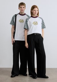 Δύο μοντέλα φοράνε λευκά T-shirts με μανίκια κοντά σε μοτίβο, που φέρουν γραφικό λογότυπο. Τους συνδυάζουν με μαύρο παντελόνι φαρδιάς γραμμής και παπούτσια.