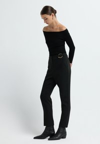 Zwarte ribbelige off-shoulder top, hoge zwarte broek met een gouden circulaire gesp, en zwarte enkellaarsjes, afgebeeld tegen een neutrale achtergrond.