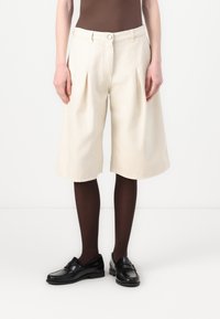 Geplooide, knielange shorts van crèmekleurige stof met een soepele textuur. Gecombineerd met zwarte instappers en donkerbruine panty’s.