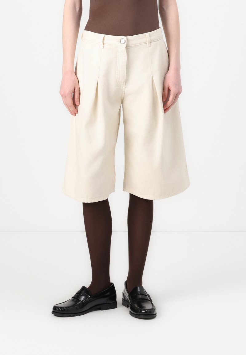 Geplooide, knielange shorts van crèmekleurige stof met een soepele textuur. Gecombineerd met zwarte instappers en donkerbruine panty’s.