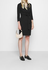 Svart omslagsklänning med trekvartsärmar, knytning i midjan, slät tyg. Kombinerad med en krämfärgad handväska och svarta loafers. Mjuk scarf runt halsen.