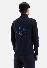 Pull en tricot bleu marine avec un col haut, doté d'accents texturés et d'un design graphique en bleu au dos. Coupe ajustée.