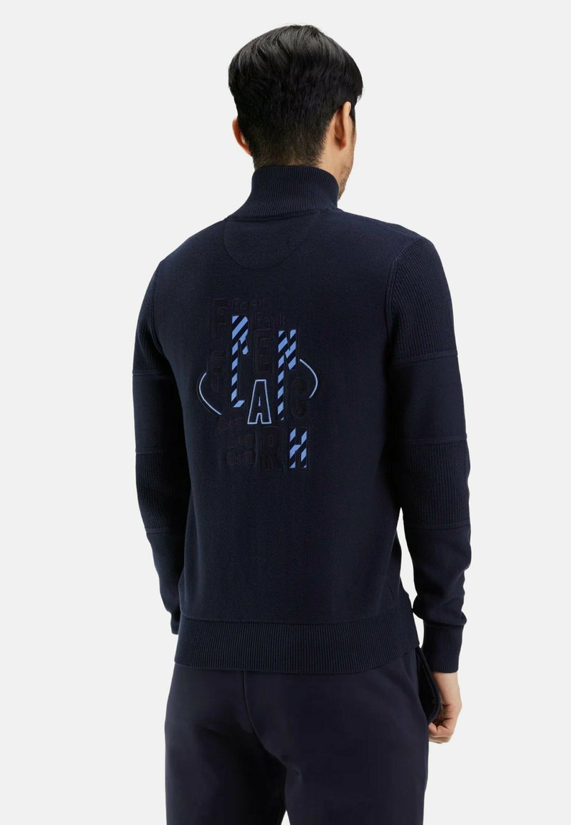 Pull en tricot bleu marine avec un col haut, doté d'accents texturés et d'un design graphique en bleu au dos. Coupe ajustée.