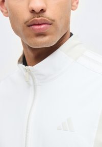 Κοντινό πλάνο του κάτω μέρους του προσώπου και του λαιμού ενός άντρα που φοράει λευκό μπουφάν με φερμουάρ και ένα διακριτικό, ελαφρύ λογότυπο Adidas στο στήθος.