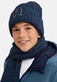 Marinblå stickad mössa med en broderad "R"-logotyp, tillsammans med en matchande scarf. Tyget visar en ribbad struktur. Modellen bär en blå jacka.