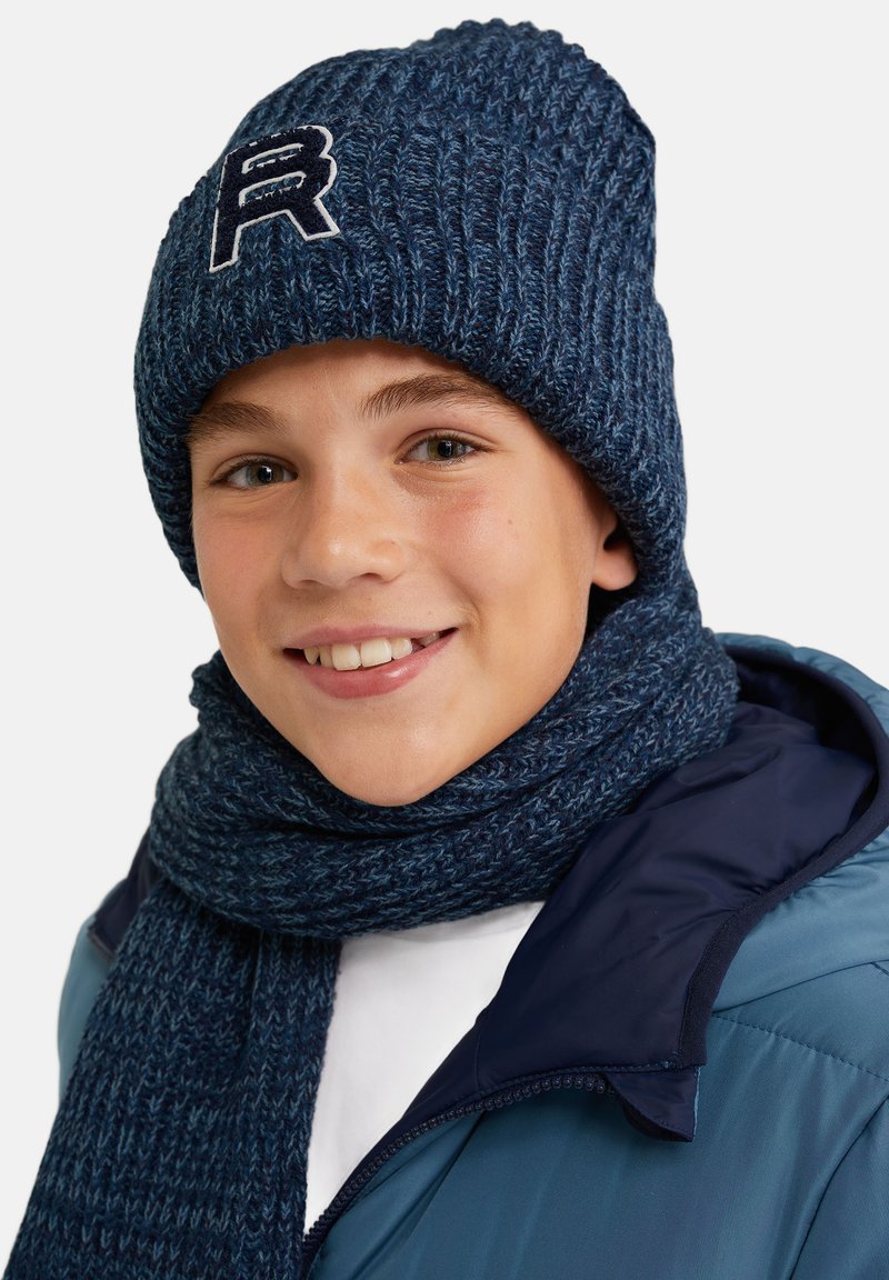 Marinblå stickad mössa med en broderad "R"-logotyp, tillsammans med en matchande scarf. Tyget visar en ribbad struktur. Modellen bär en blå jacka.