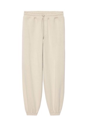 Joven con el pelo corto que lleva una camisa blanca de manga larga, pantalones deportivos beige y zapatillas blancas, de pie con las manos en los bolsillos.