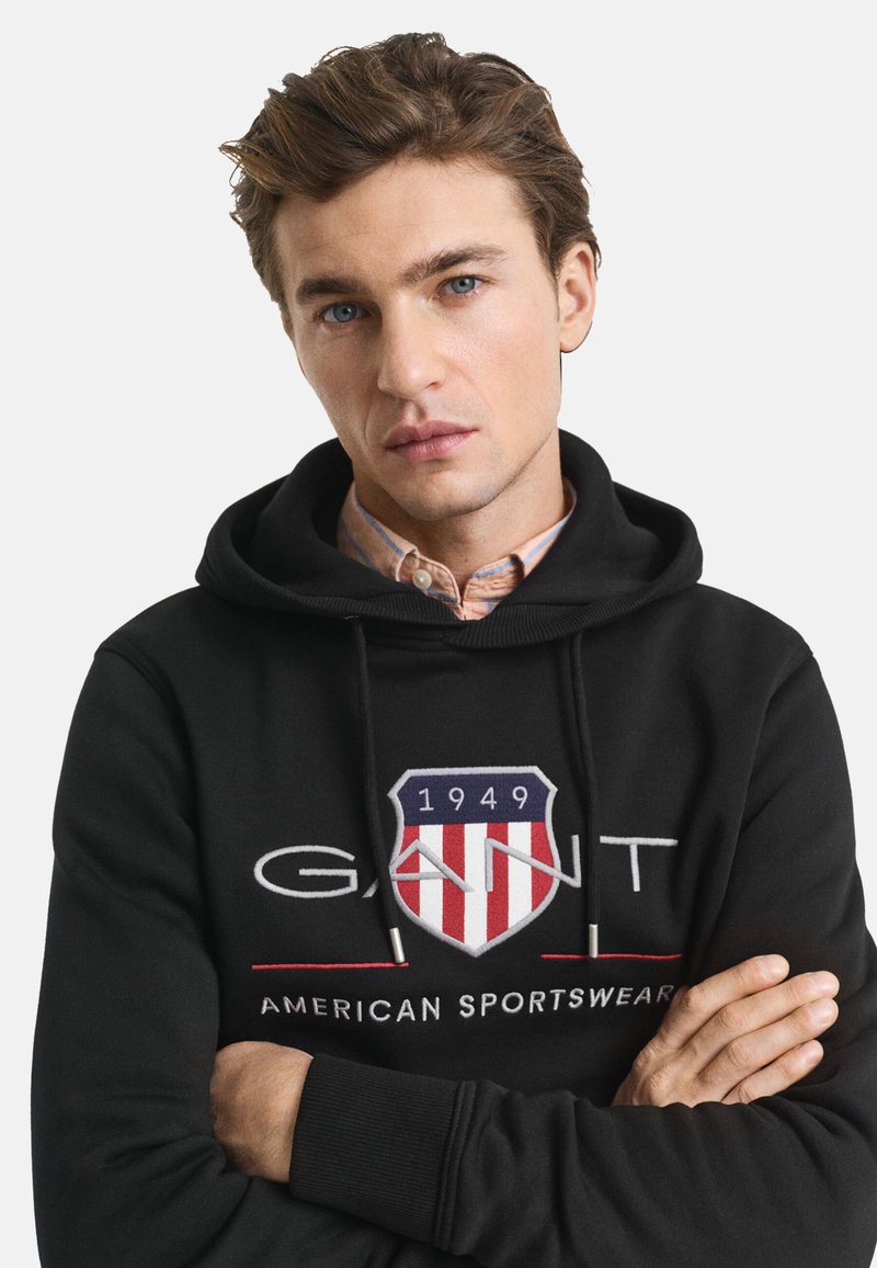 Svart hoodie i mjuk bomull med dragsko. Har en framträdande GANT-logotyp med skölddesign och röd, vit och blå accent.