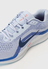 Chaussure de course Nike gris clair et bleu avec tige en mesh, logo swoosh bleu marine, lacets blancs et semelle coussinée avec texte rouge "Air".
