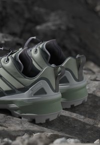 Zielone i szare buty sportowe z teksturowaną powierzchnią, wyposażone w solidną podeszwę, tylną pętelkę do zakupu i wyraźne oznaczenie marki z boku.
