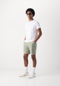 Mannenoutfit met een witte short-sleeve t-shirt, lichtgroene shorts, witte sneakers met bruine accenten en zwarte zonnebril.