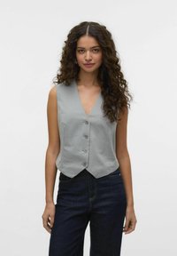 Grå ärmlös väst med V-hals, fem knappar och en figurnära design, i kombination med mörkblå jeans. Slät tygtextur.