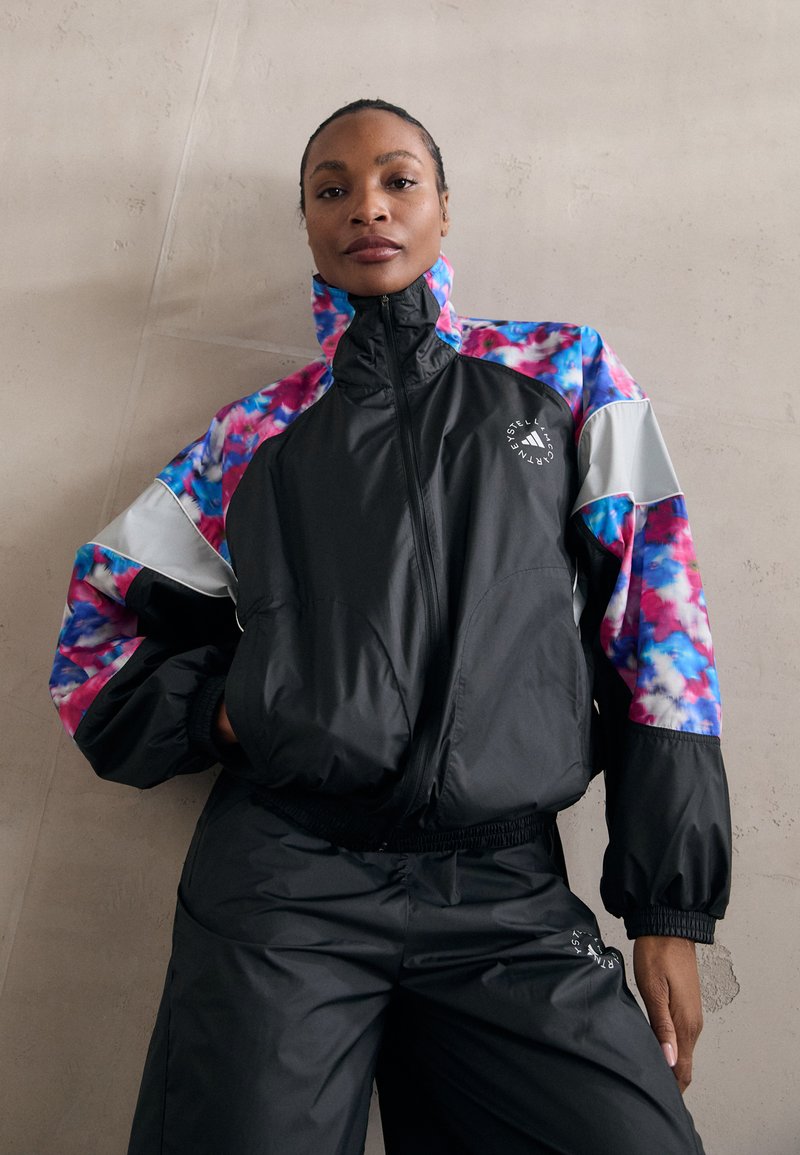 Veste noire légère avec des manches colorées tie-dye. Elle présente un col haut, un devant zippé et des poignets élastiques. Texture lisse.