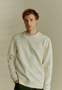 Felpa crema con un design testurizzato a motivo diamante, colletto rotondo e polsini a costine, abbinata a pantaloni verde scuro.