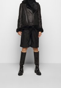 Svart läderjacka med pälskrage, midjebälte och långa ärmar. Kombinerad med svarta läderbyxor och glänsande knähöga stövlar.