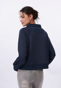 Mørkeblå cropped hettegenser med høy krage og ribbekanter. Glatt stoff; har en bakstykkedetalj og en avslappet passform. Kombinert med lysegrå jeans.