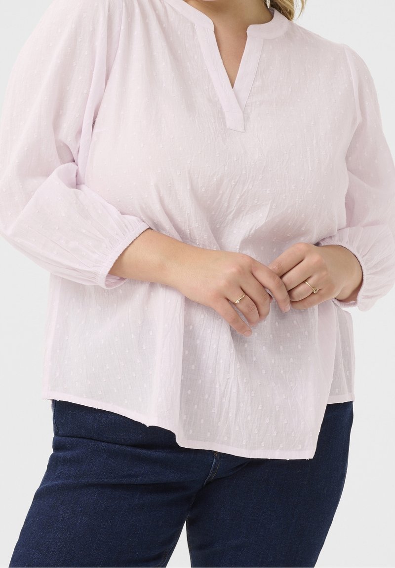 Jasnoróżowa bluzka o fakturowanej powierzchni z dekoltem w kształcie litery V i bufkowymi rękawami. Materiał jest pokryty małymi kropkowymi wzorami, zestawiony z ciemnoniebieskimi dżinsami.
