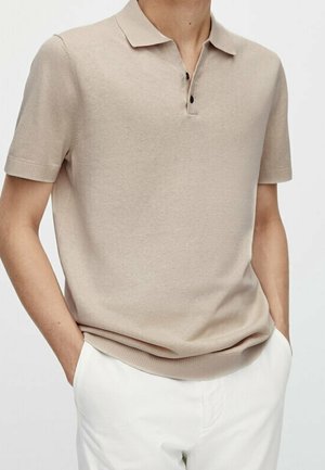 Man wearing a beige short-sleeve polo shirt with black buttons and white pants, hands in pockets, against a plain background.