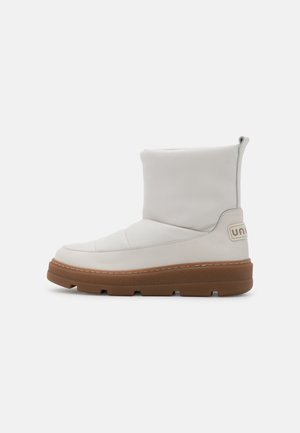 Bottes blanches à cheville en matériau lisse, design rembourré, et semelle en caoutchouc marron clair. Dotées d'une boucle à l'arrière et de détails cousus.