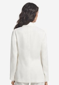 Vit kavaj gjord av ett mjukt, lätt tyg. Har en enda söm baktill, klövd krage och långa ärmar med en ren avslutning.