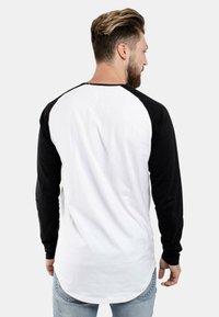 Langarm-Baseballshirt mit einem weißen Körper und schwarzen Ärmeln. Verfügt über einen abgerundeten Saum und ein dezentes Logo auf der Rückseite. Baumwollmaterial.
