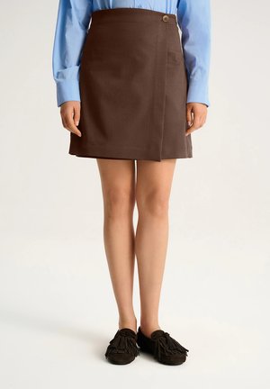 Kobieta ubrana w brązową spódnicę zapinaną z guzikiem i czarne mokasyny z frędzlami, zestawione z jasnoniebieską koszulą z długim rękawem.