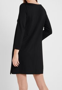 Robe noire en A avec manches longues, col rond et texture lisse. Présente une fente sur le côté pour faciliter le mouvement et des détails de couture minimaux.
