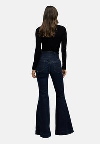 Vaqueros de campana de talle alto en denim oscuro con un acabado suave. Presenta dos bolsillos traseros y sutiles detalles de costura.