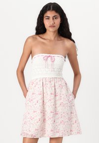 Axelös klänning med en virkade vit överdel och en kjol med blommigt rosa mönster, utrustad med fickor, knuten med ett rosa dragsnöre.