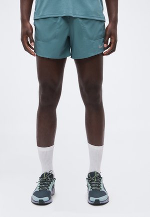 Man met blauwgroene sportshorts, lichtblauwe en zwarte hardloopschoenen en witte sportsokken, staand tegen een effen witte achtergrond.